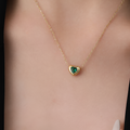 A delicate gold chain necklace with a small heart-shaped pendant featuring a green heart design in the center, shown worn on a model's neck.
